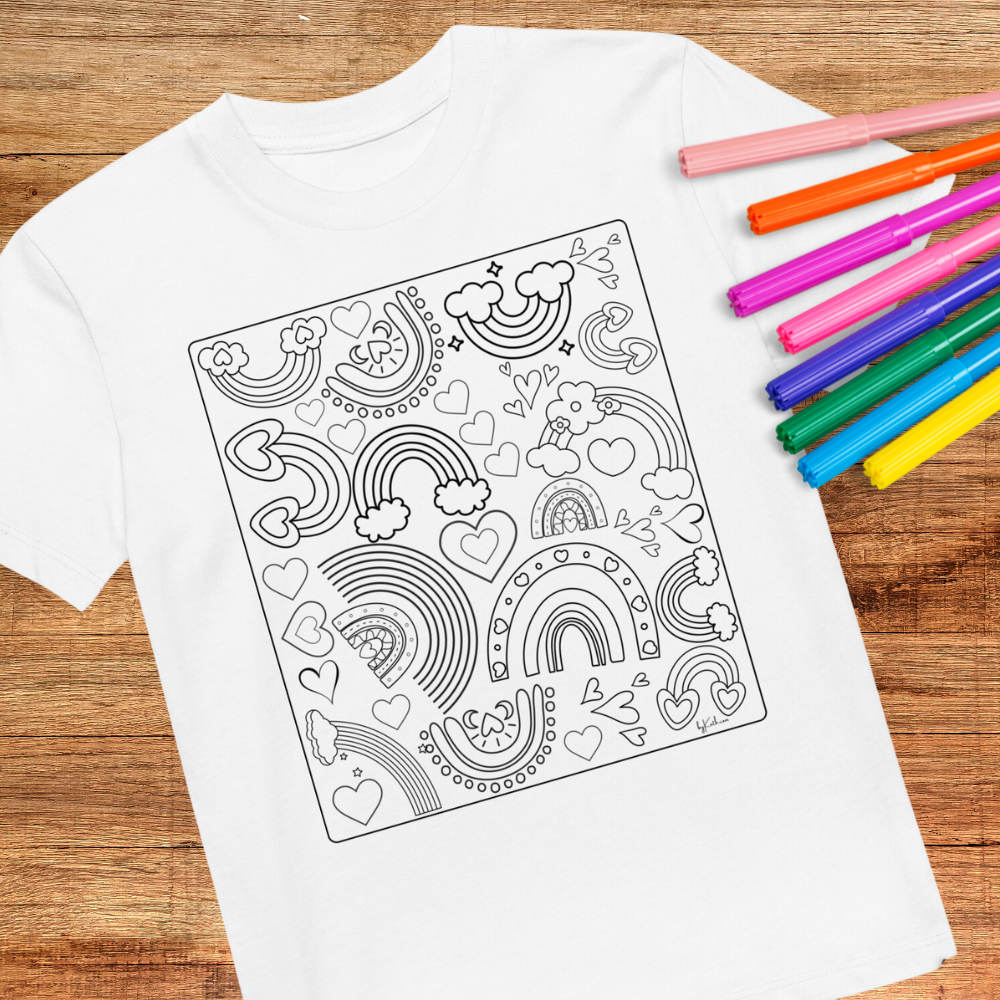 A Rainbow Doodles Color-in tee: Organic cotton kids t-shirt by byKath.com, featuring a black and white coloring design with rainbows, hearts, clouds, and stars, is laid flat on a wooden surface. To the right of the shirt are seven colorful markers in shades of orange, pink, purple, blue, and green—perfect for an interactive coloring experience.