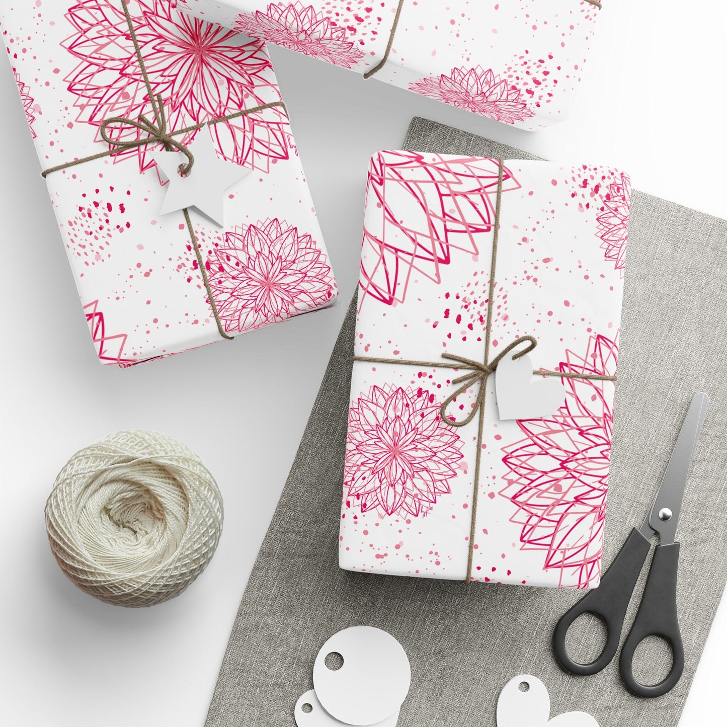 A neatly wrapped gift in byKath's Designed byKath.com Pink and white floral Wrapping Paper, tied with twine and a heart-shaped tag. Surrounding the gift are a ball of twine, a pair of scissors, and several blank tags, all placed on a gray fabric surface.