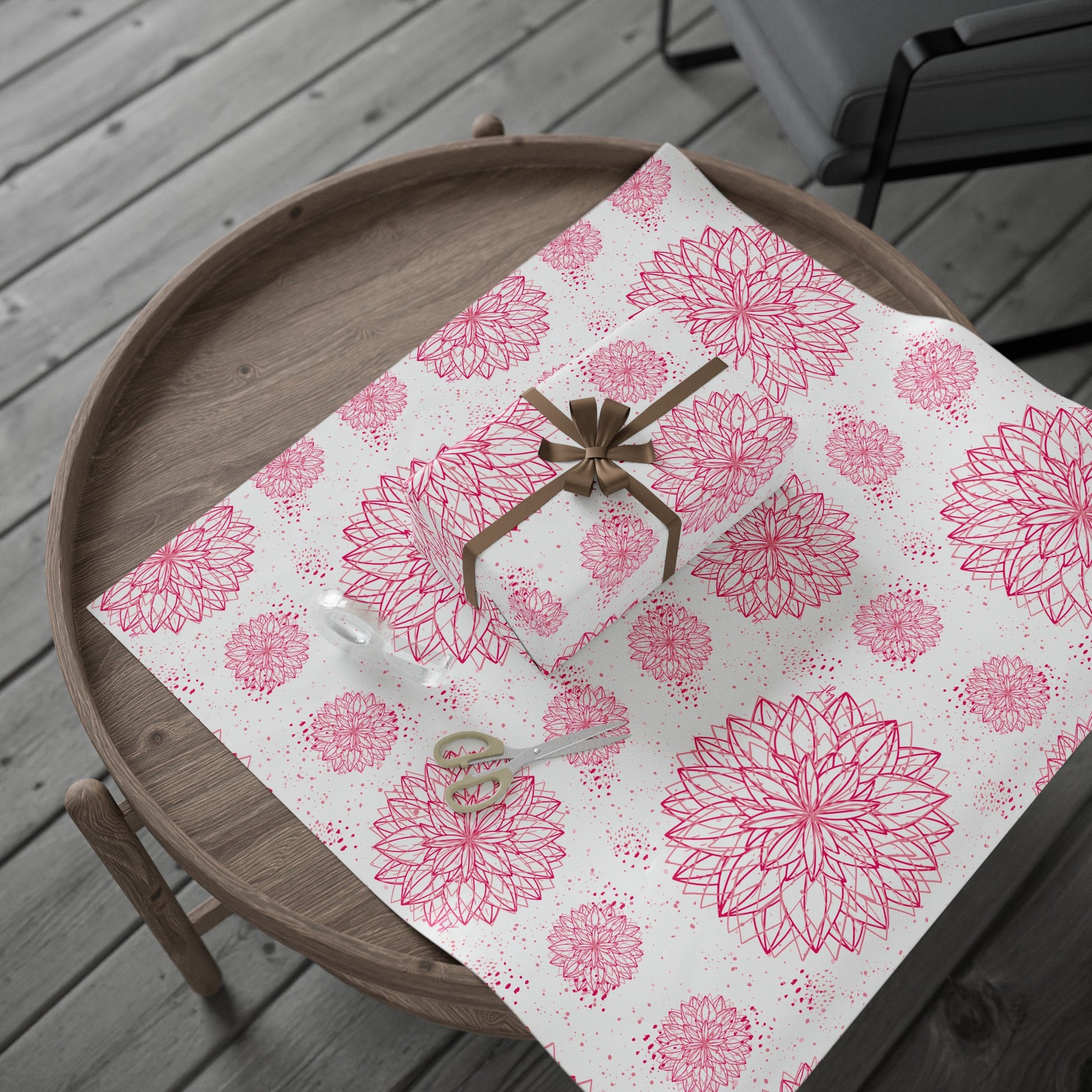 A wooden round coffee table showcasing a beautifully wrapped gift in byKath's Designed byKath.com Pink and White Floral Wrapping Paper, adorned with a gold ribbon. Alongside the gift, you'll find a pair of scissors and a piece of clear tape on a dispenser. The entire setup rests on matching high-definition print wrapping paper from the same collection. In the background, there's a wooden floor adding to the aesthetic charm.