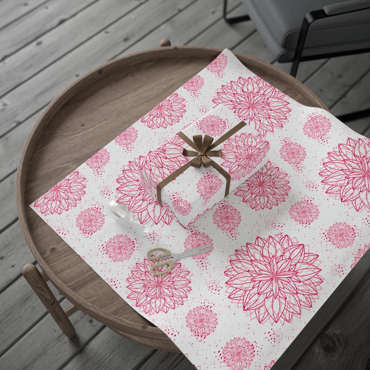 A round wooden table is set with byKath’s Designed byKath.com Pink and white floral Wrapping Paper in various sizes, showcasing its beautiful pink floral patterns. A gift box wrapped in the same high-definition print and tied with a golden ribbon sits on top. A pair of scissors and a roll of clear tape lie next to the gift on the paper.