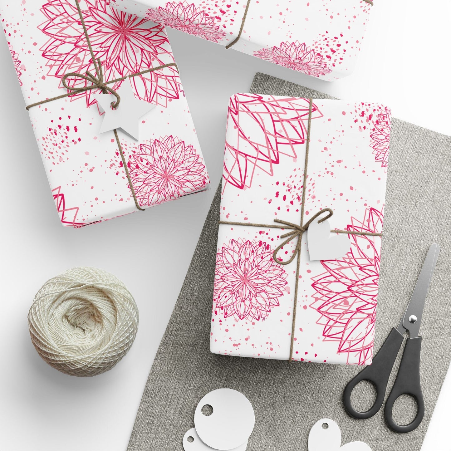 Beautifully wrapped gifts in elegant pink and white floral designs, printed in high definition on byKath’s fine-art wrapping paper, are tied with twine and blank tags. The scene includes a roll of twine, scissors, and blank gift tags, all placed on a neutral-colored surface. The wrapping paper is from Designed byKath.com and is available in various sizes.