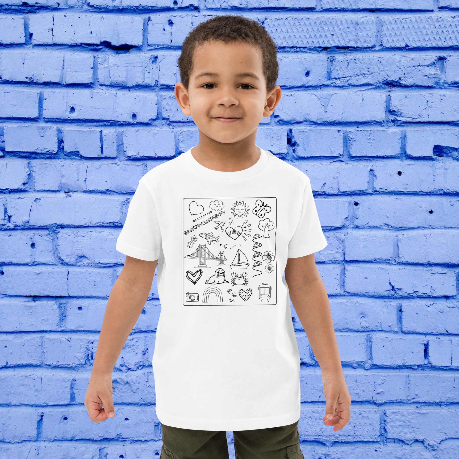 A young child with short hair is standing and smiling against a blue brick wall. The child is wearing an oversized white T-shirt featuring various illustrated doodles from the San Francisco Color-in Tee: Organic cotton kids t-shirt collection by byKath.com, paired with khaki pants.