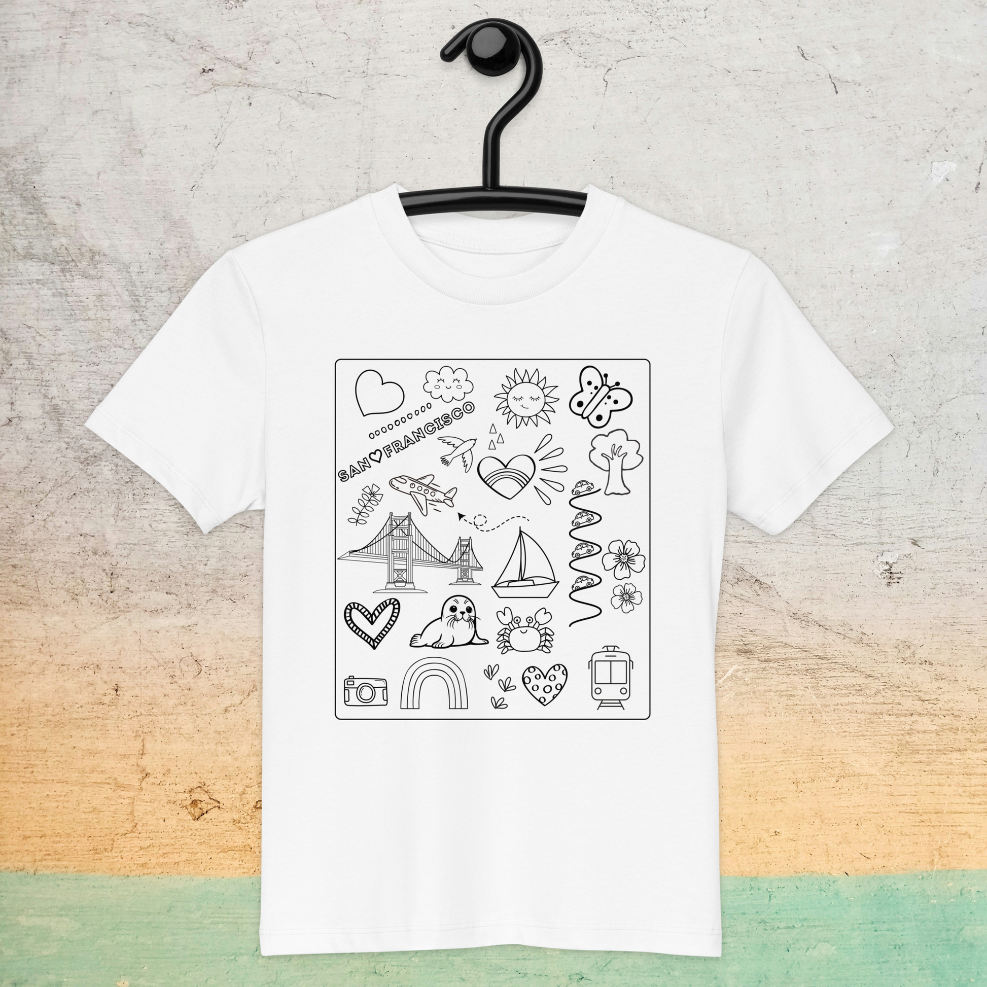 A white San Francisco Color-in Tee: Organic cotton kids t-shirt by byKath.com hangs on a black hanger against a textured background. The shirt features black line art drawings, including a rainbow, lighthouse, Eiffel Tower, boat, dog, butterfly, sun, cloud, and various other playful doodles.