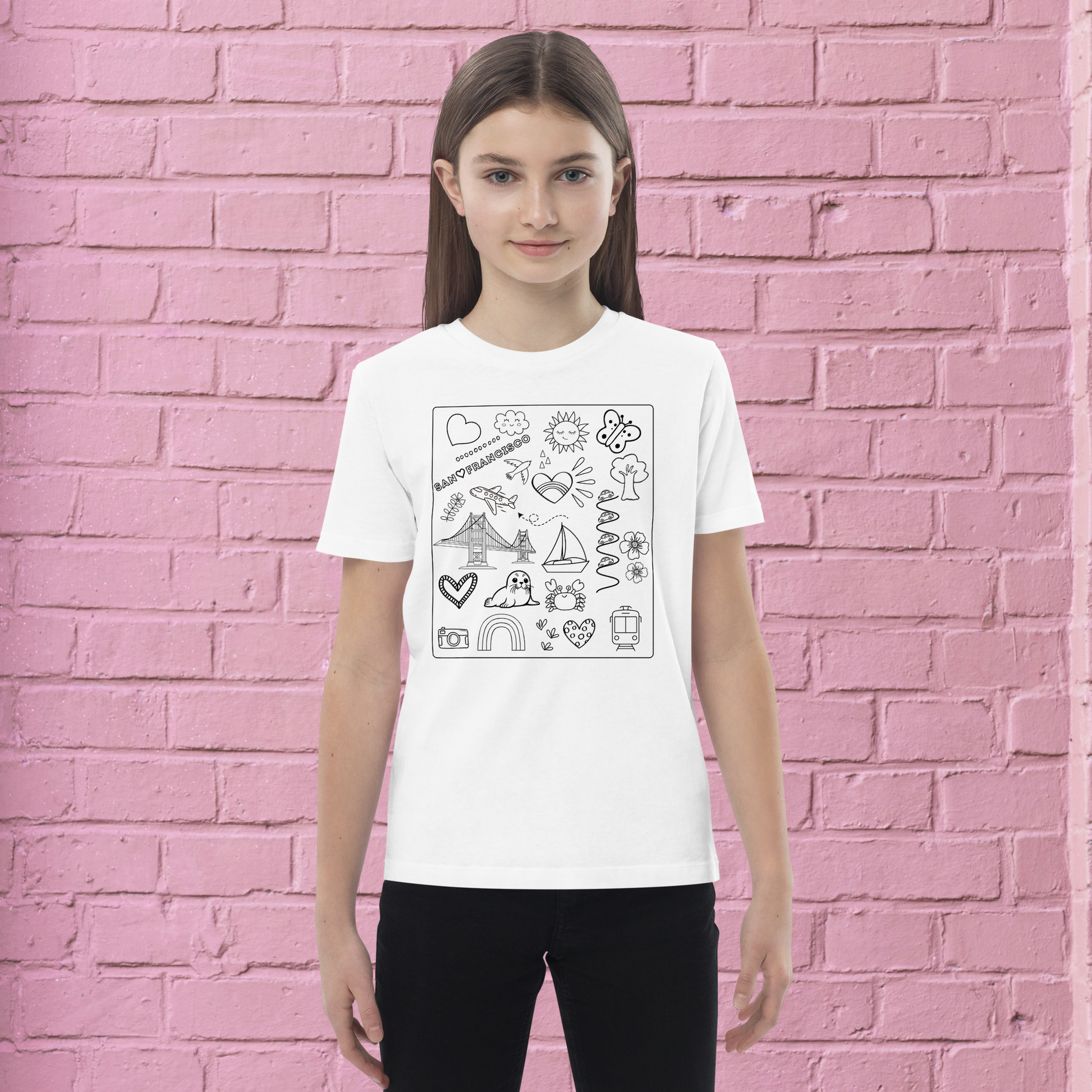 A young person with long brown hair stands against a pink brick wall, sporting the San Francisco Color-in Tee from byKath.com. The shirt, designed for kids and made of organic cotton, features black outline illustrations including hearts, a rainbow, and clouds. This piece of interactive kids' clothing provides both comfort and a canvas for creativity. Their arms are relaxed at their sides with a neutral expression on their face.