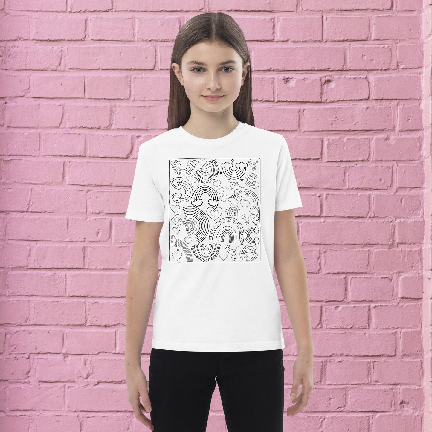 A young person with long hair and a white Rainbow Doodles Color-in tee by byKath.com stands in front of a pink brick wall. The organic cotton kids' T-shirt showcases a black and white abstract design featuring rainbows, fruits, and various shapes, making it perfect for an interactive coloring experience. The person gazes directly at the camera with a neutral expression.