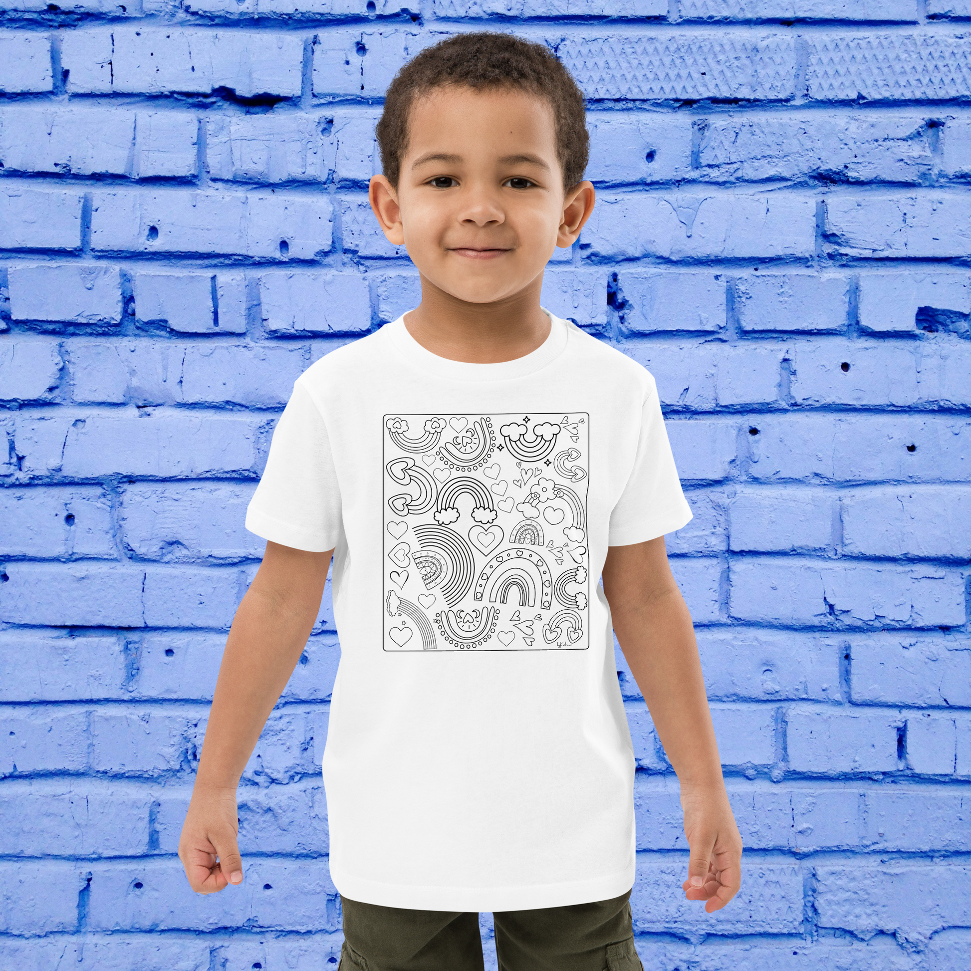 A young child stands smiling against a blue brick wall, wearing the Rainbow Doodles Color-in tee from byKath.com. The white organic cotton kids' t-shirt features a black and white design with abstract patterns, including rainbows, clouds, and various shapes. This customizable gift idea pairs well with his short, dark hair and olive green shorts.