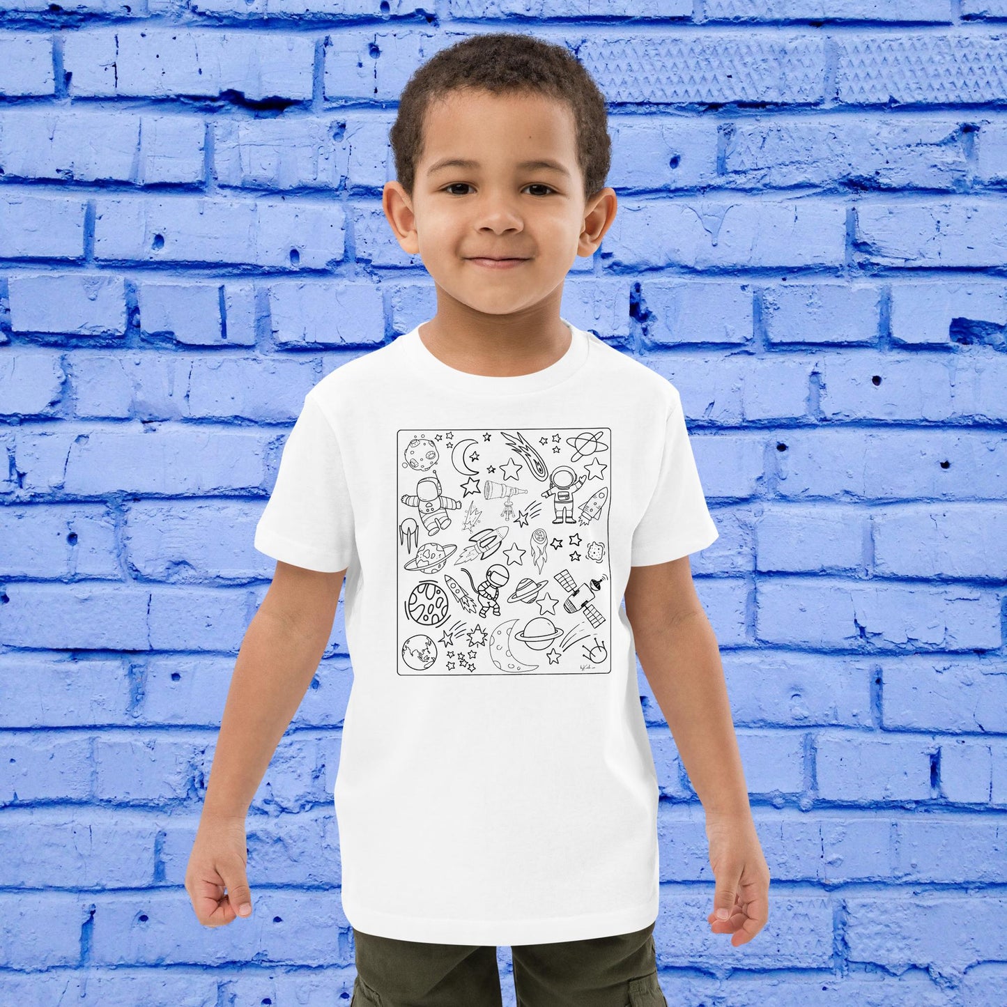 A young boy stands smiling against a bright blue brick wall, wearing a Space Adventure Doodles Color-in tee: Organic cotton kids t-shirt by byKath.com that features a black and white doodle design of various space-themed illustrations like rockets, planets, and stars. Dressed in green pants, he’s ready for some Space-Themed Coloring Fun.