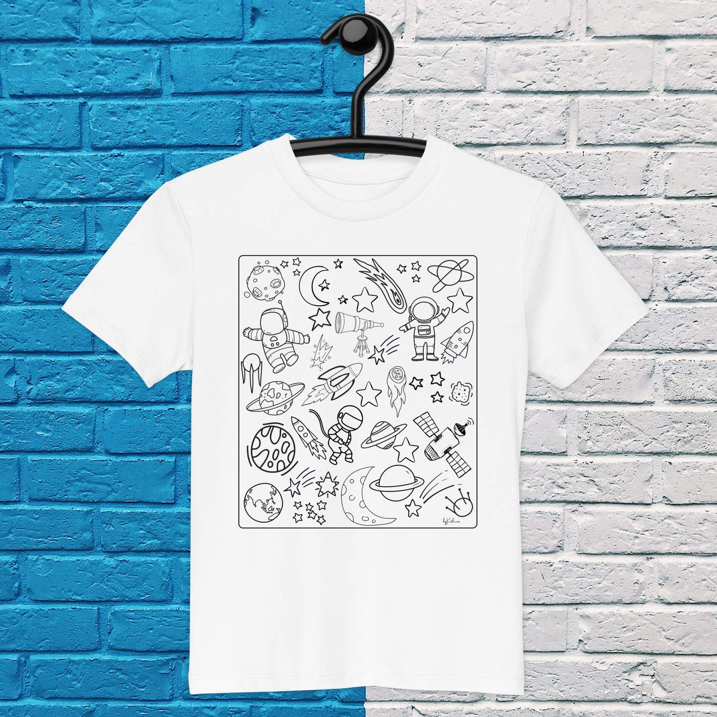 A white Space Adventure Doodles Color-in Tee: Organic Cotton Kids T-Shirt by byKath.com, featuring black line art drawings of astronauts, planets, rockets, stars, satellites, and space stations, is displayed hanging on a hook against a two-tone blue and white brick wall background. This space-themed coloring shirt is perfect for young explorers.