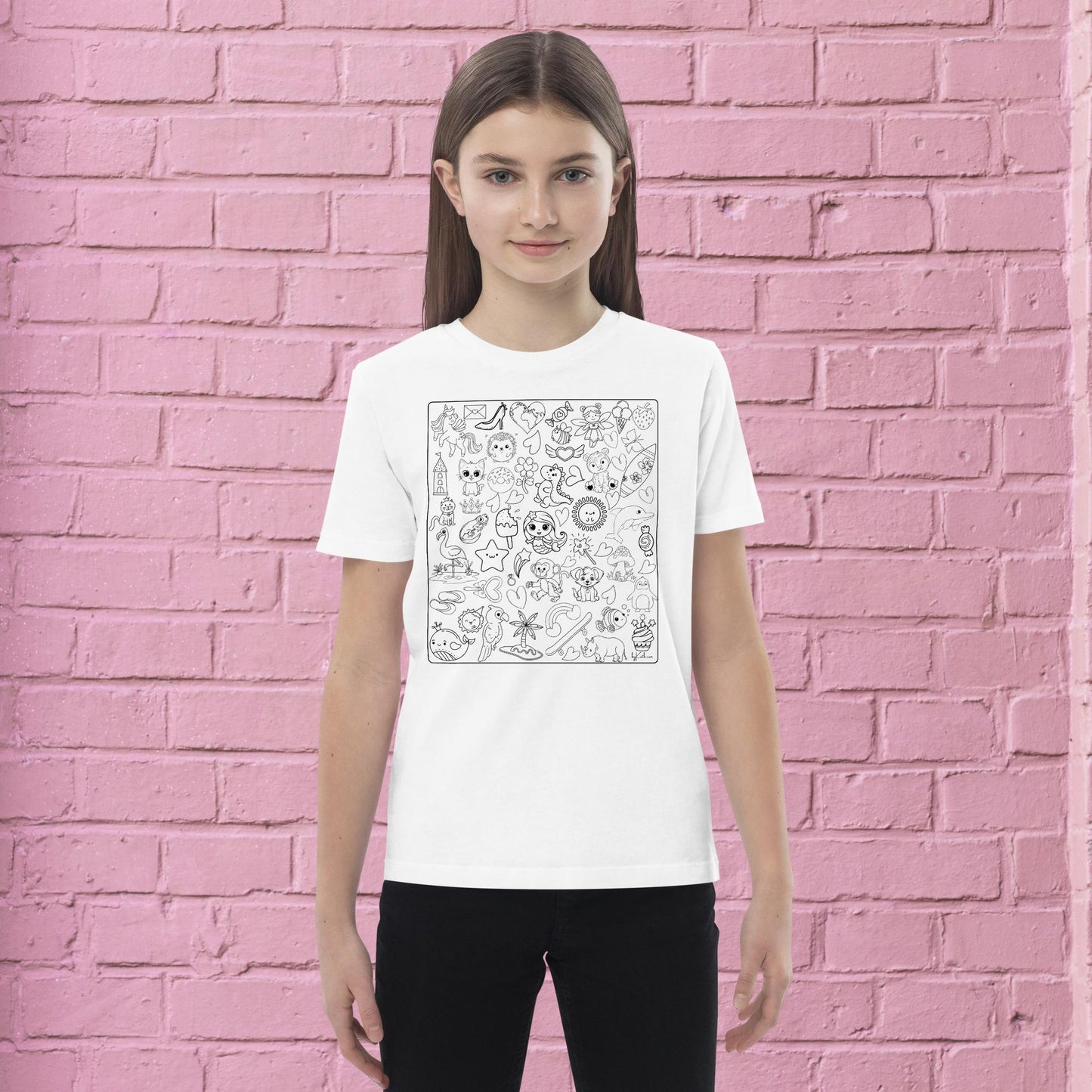 A person with long hair stands against a pink brick wall, wearing a white Happy Doodles Color-in Tee: Organic cotton kids t-shirt by byKath.com adorned with a square design showcasing an array of detailed, whimsical sketches. The person is smiling slightly and has their hands by their sides in mindful moments. They are also wearing black pants.