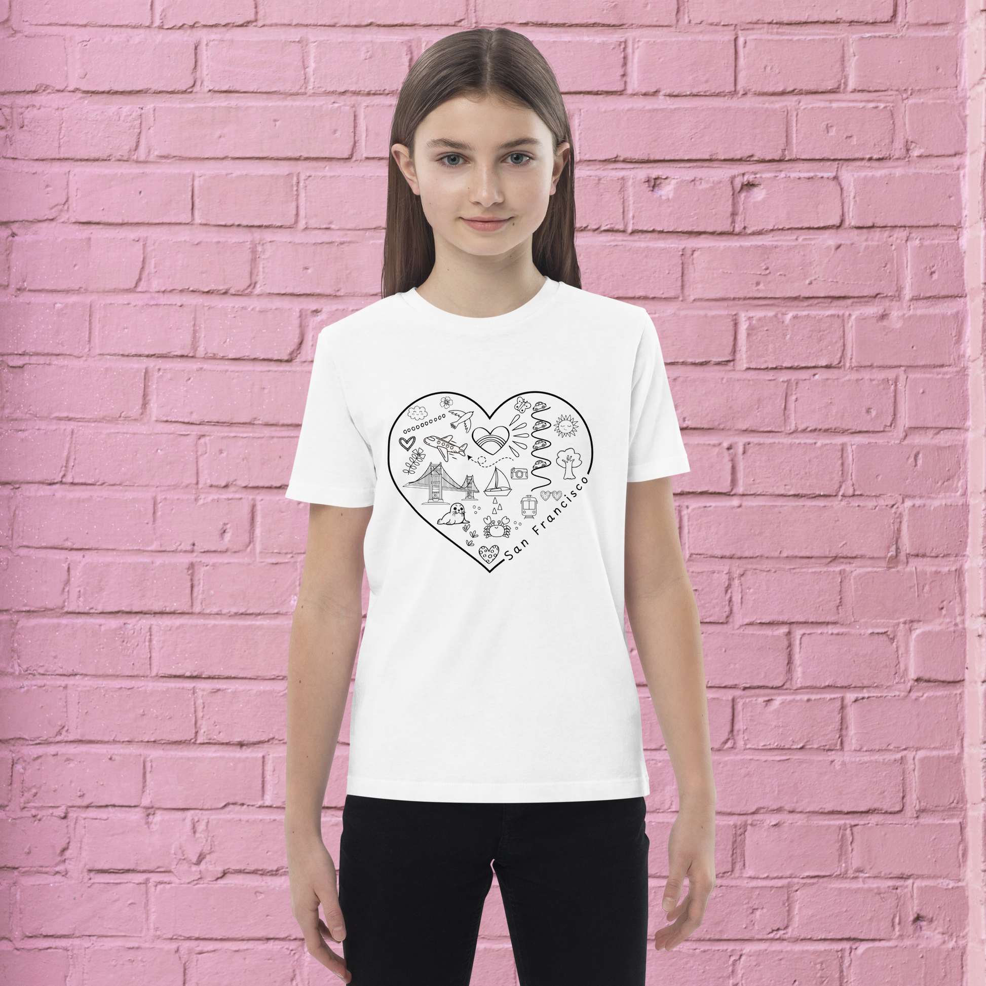 A young person stands against a pink brick wall, wearing a San Francisco Color-in Tee: Organic cotton kids t-shirt by byKath.com. This white T-shirt features a heart-shaped design with various San Francisco landmarks and icons, making it perfect for creative exploration.