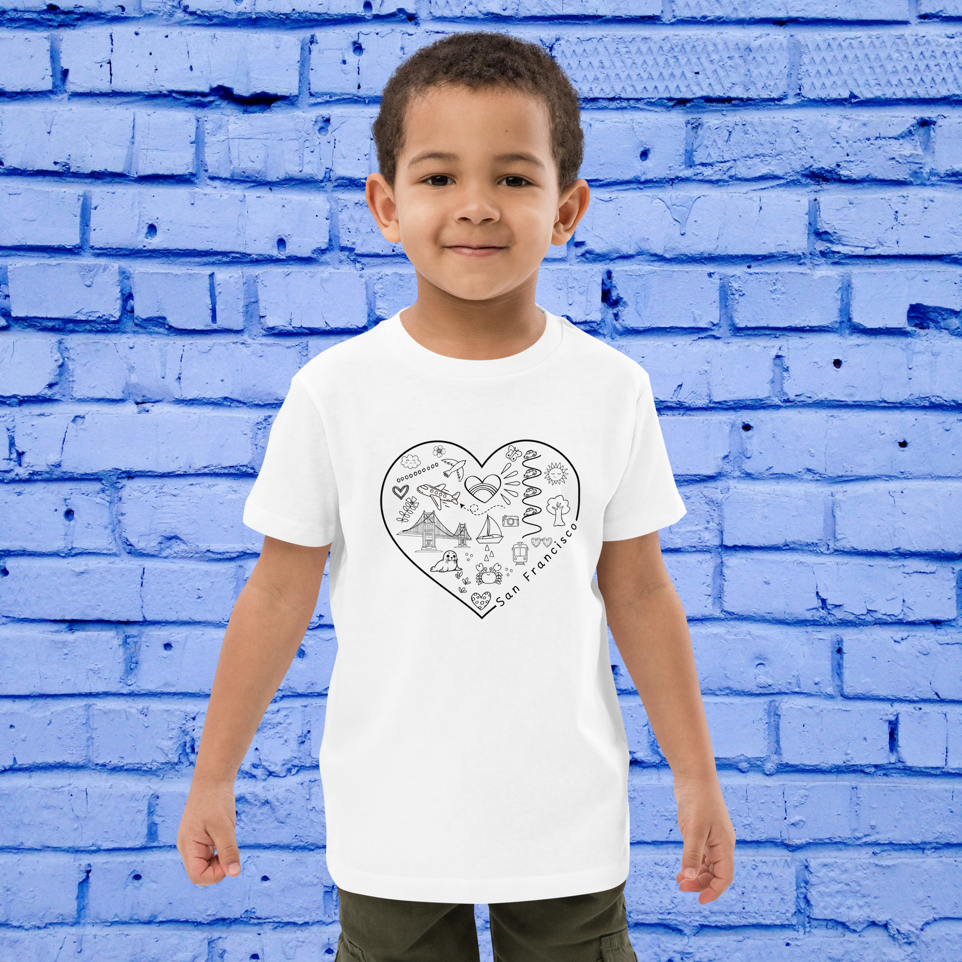 A young child stands in front of a blue brick wall, wearing a San Francisco Color-in Tee by byKath.com. This organic cotton kids' t-shirt is adorned with heart-shaped San Francisco-themed illustrations. The friendly child stands with hands at their sides, highlighting the playful charm of interactive kids' clothing.